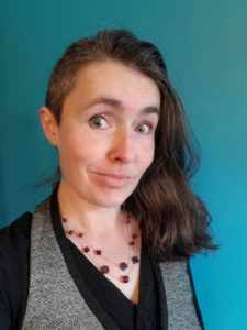 A dark haired woman smirks at the camera, her eyebrows high. She wears a grey vest, black shirt, red stone necklace, and stands in front of a teal blue backdrop.