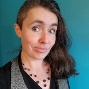 A dark haired woman smirks at the camera, her eyebrows high. She wears a grey vest, black shirt, red stone necklace, and stands in front of a teal blue backdrop.